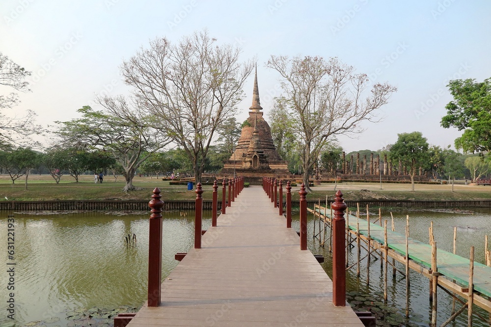 Fototapeta premium Scenery of Wat Sa Si, an ancient Buddhist Temple in Sukhothai Historical Park, with the majestic Pagoda (Stupa) in background & a wooden bridge across the canal ~ A beautiful heritage site in Thailand