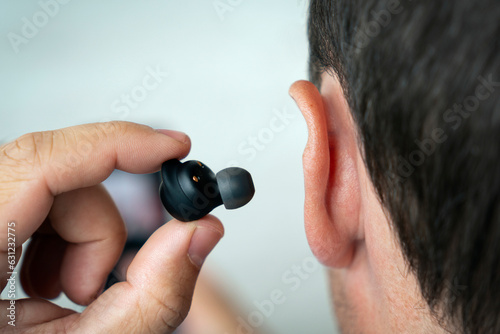 Photos Sideways portrait of smiling young man listening to music or radio, uses modern wireless earphones, wearing blue t-shirt