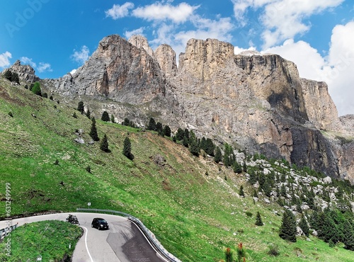 Magnificent scenery of Dolomiti under blue sky with rugged Sella group in background & cars traveling on a sharp turn of a mountain highway by mountainside in Passo Sella, Trentino, South Tyrol, Italy photo