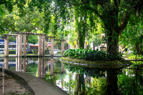 Captivating Reflections  Vibrant Greenery and Salmon-Colored Monument Mirror on Tranquil Lake. Serene scene captures bustling avenue  passing buses  and vibrant nature amidst urban oasis.