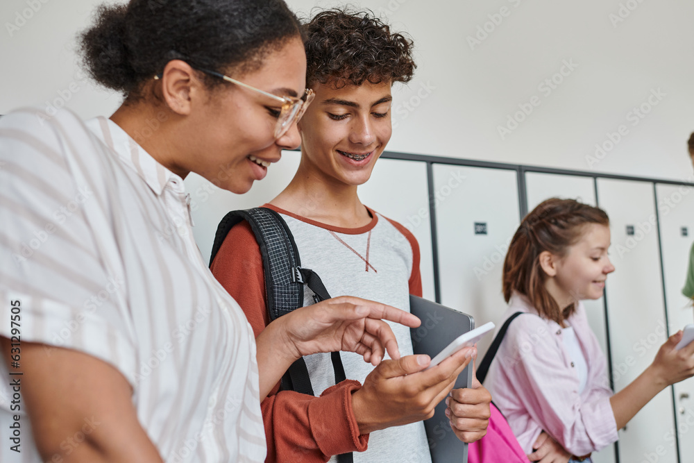 Fototapeta premium happy african american teacher pointing at smartphone of student, teenagers in school hallway