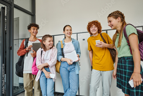 cheerful teenage students holding devices and chatting in school hallway, teen friends, student life