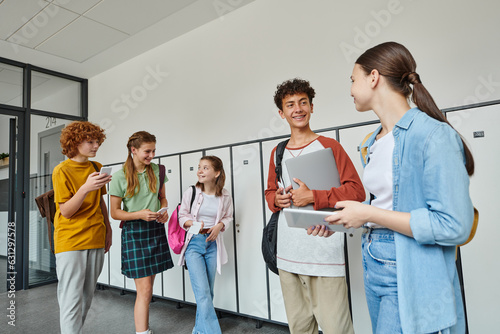 happy classmates talking and holding devices in hallway, back to school, teen students, digital age