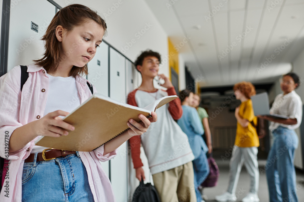 schoolgirl holding notebook and standing near blurred and happy teen classmate, students, hallway