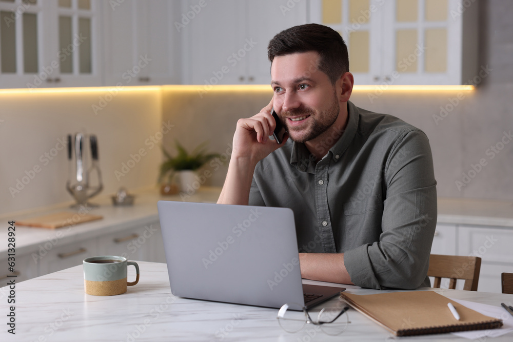 Happy man talking on phone while working with laptop at white marble table in kitchen Happy man talking on phone while working with laptop at white marble table in kitchen