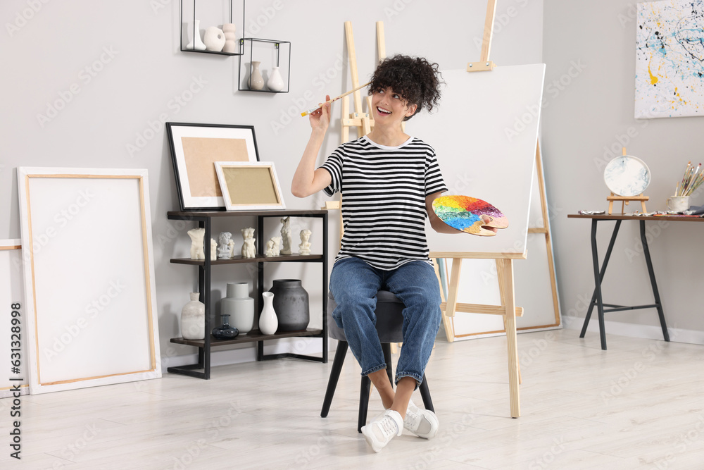 Young woman with brush and artist`s palette near easel in studio Young woman with brush and artist`s palette near easel in studio