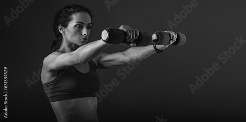 Female sporty muscular young serious woman doing strength workout on the shoulders, biceps and arms in sport bra holding dumbbells on black background with empty space. Closeup