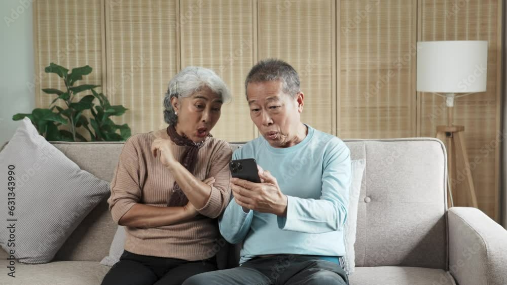 Middle-aged Aaian couple using smartphone together or making video call while sitting at sofa in cozy living room. Middle-aged Aaian couple using smartphone together or making video call while sitting at sofa in cozy living room.