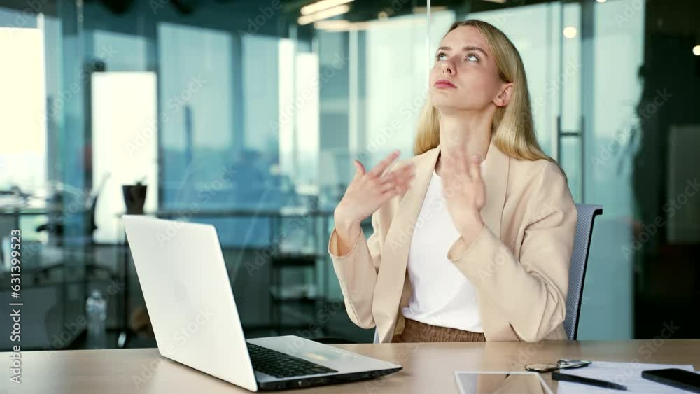 A young businesswoman is hot sitting at the workplace at the desk in a modern office. Woman waves her hands like a fan, cannot concentrate on work, because the air conditioner in the room is broken