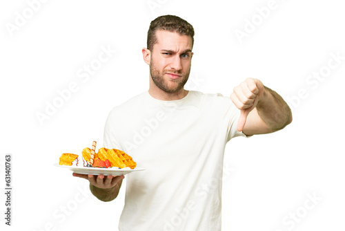Young handsome blonde man holding waffles over isolated chroma key background showing thumb down with negative expression
