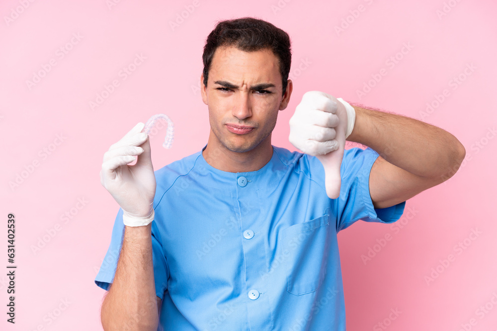 Dentist caucasian man holding invisible braces isolated on pink background showing thumb down with negative expression Dentist caucasian man holding invisible braces isolated on pink background showing thumb down with negative expression
