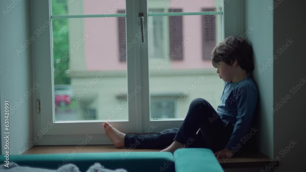Bored child sitting by window looking at drizzle fall during rainy afternoon. Little boy stuck at home wanting to go out, sad thoughtful expression