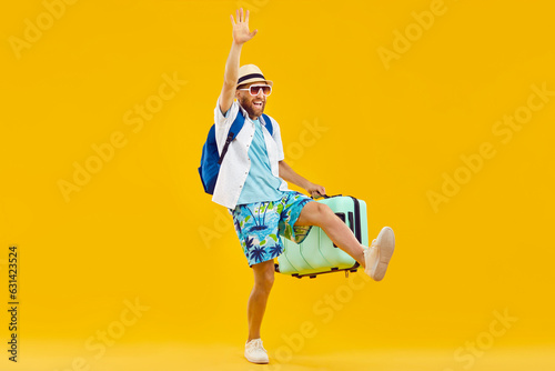 Joyful happy man with suitcase is delighted to go on summer vacation to sea. Caucasian millennial man in summer clothes holding suitcase waves and laughs against orange background. Full length. Banner photo