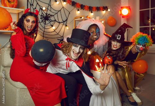 Group of friends disguised as crazy evil monsters having fun at Halloween party. Adult people with scary makeup sitting together on sofa in living room, looking at camera, smiling or making ugly faces