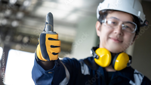 Confident Asian man construction worker with uniform suit, ear muffs, safety helmet and goggles showing thumbs up signal in protective gloves. Male engineer showing hand gesture at construction site photo