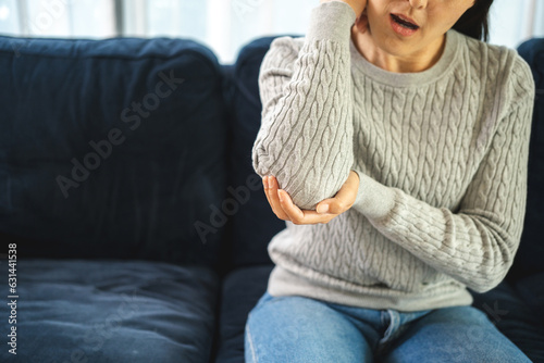 Young woman has problem with structural posture arms elbow shoulder pain. Massage her elbow and shoulders for relief. reduce muscle tension on sofa couch in living room