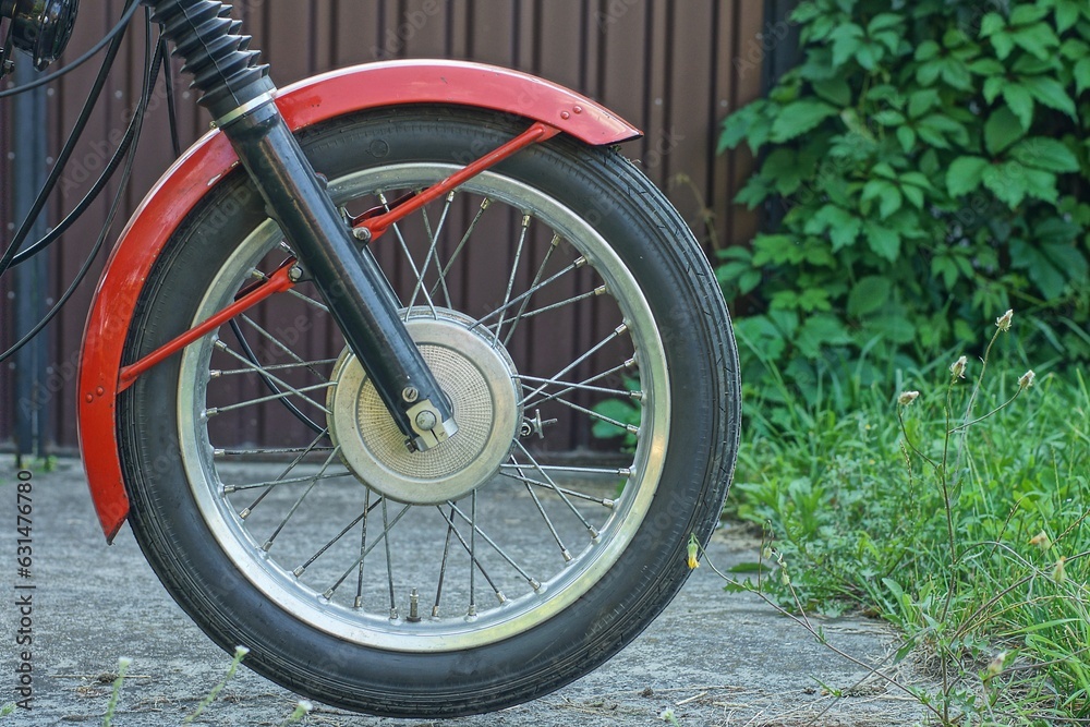 one front old retro wheel with fork part and red  color front iron fender of old classic java motorcycle outdoors on the street near brown fence
