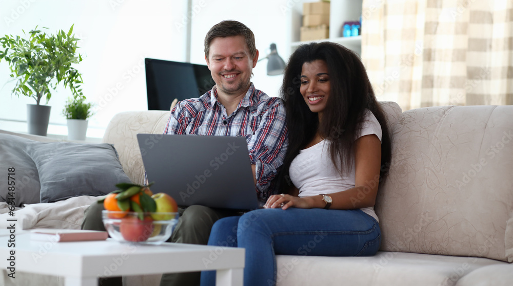 Portrait of happy smiling couple viewing photos, watching movie on laptop. Man and african-american woman sitting on cozy sofa. Home interior and mixed race marriage concept Portrait of happy smiling couple viewing photos, watching movie on laptop. Man and african-american woman sitting on cozy sofa. Home interior and mixed race marriage concept
