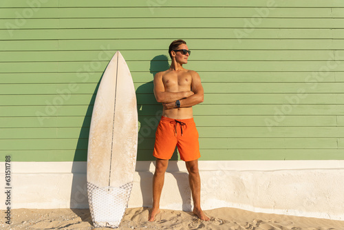 A smiling man with nice body wearing sunglasses standing at the light green with a surfboard - looking to the side photo