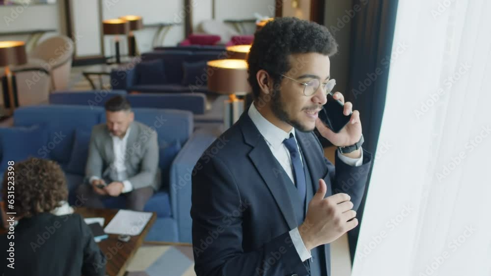 Medium shot of businessman standing next to window and talking on smartphone while spending time at business lounge at hotel with other guests Medium shot of businessman standing next to window and talking on smartphone while spending time at business lounge at hotel with other guests