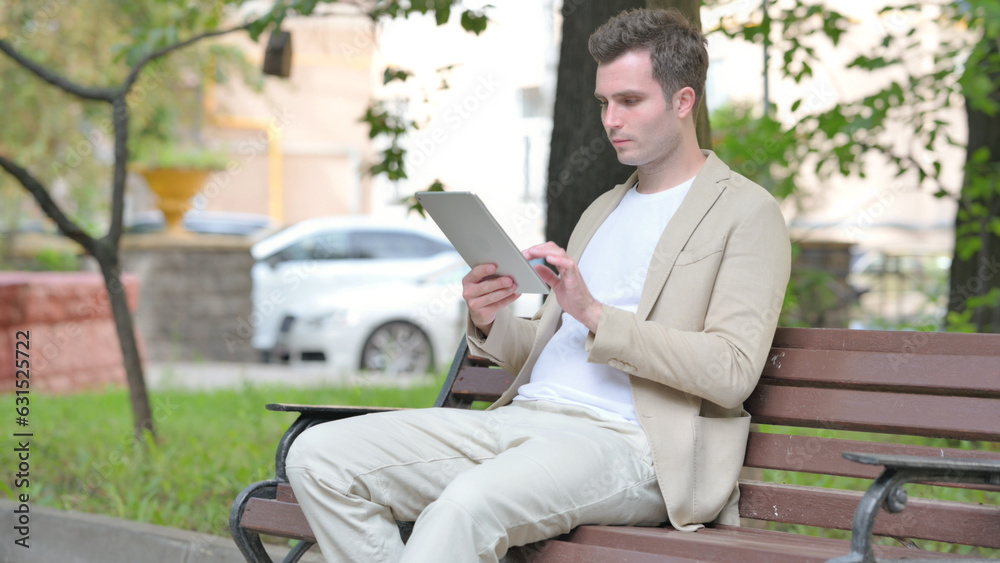 Obraz premium Casual Young Man using Tablet while Sitting Outdoor on a Bench