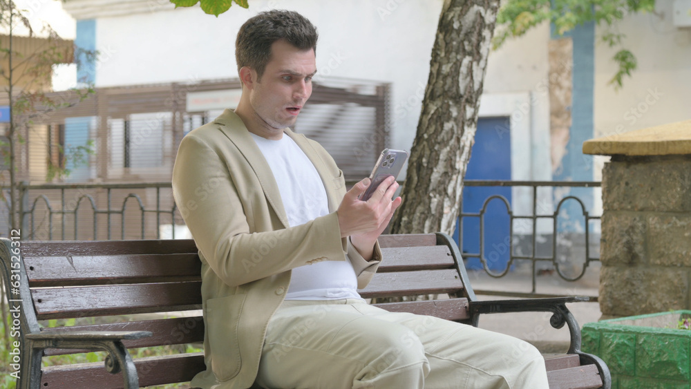 Young Man Shocked by Loss on Smartphone while Sitting Outdoor on a Bench Young Man Shocked by Loss on Smartphone while Sitting Outdoor on a Bench