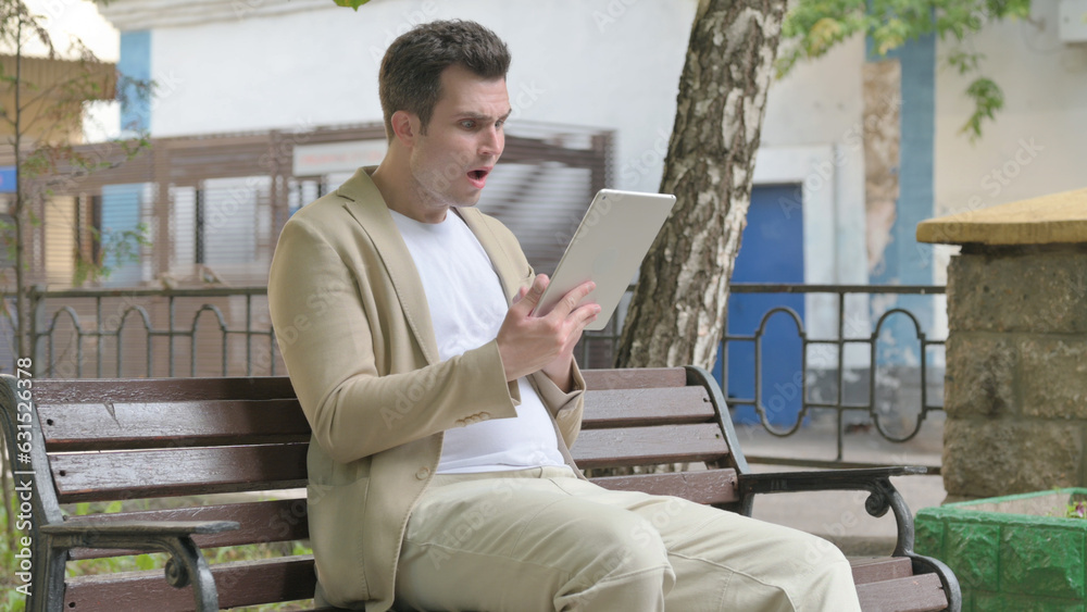 Young Man Upset by Loss on Tablet while Sitting Outdoor on a Bench Young Man Upset by Loss on Tablet while Sitting Outdoor on a Bench