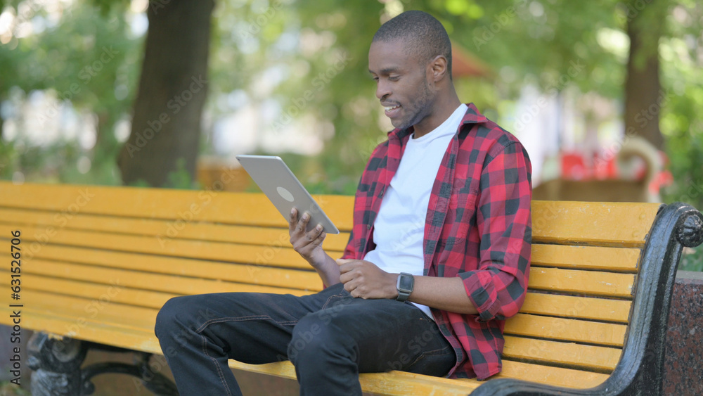 Obraz premium Young African Man Doing Video Chat on Tablet while Sitting Outdoor on a Bench