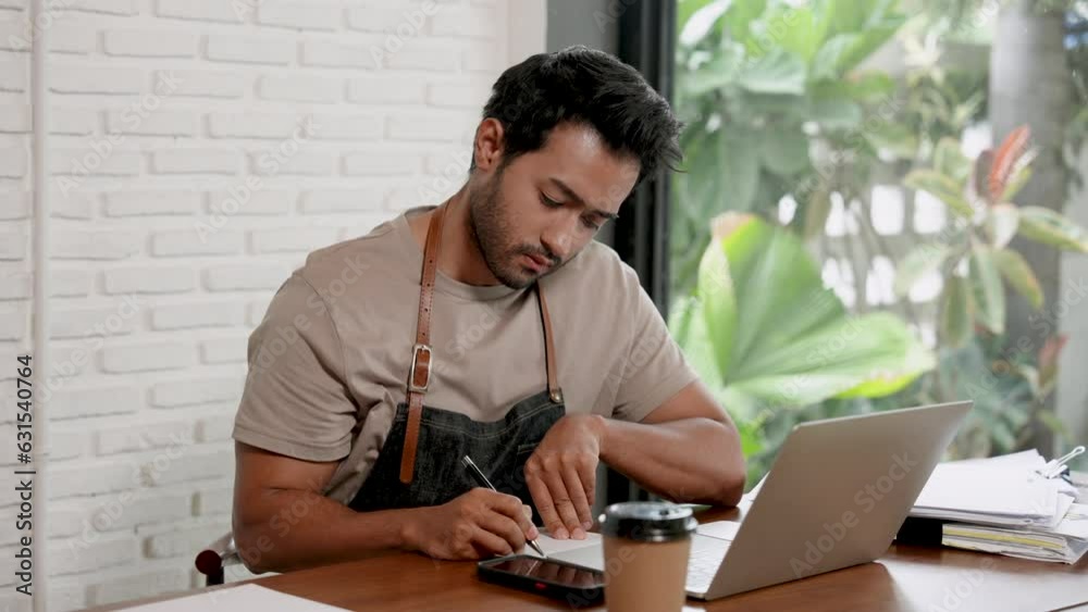Handsome Indian man is a barista and owner of small business cafe. sitting at the table take your mind seriously to prepare to order raw materials of the coffee shop via online with notebook computer Handsome Indian man is a barista and owner of small business cafe. sitting at the table take your mind seriously to prepare to order raw materials of the coffee shop via online with notebook computer