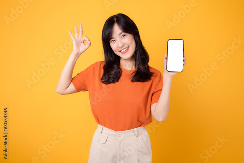 Vibrant young Asian woman in her 30s, wearing an orange shirt, shows okay gesture and smartphone blank display screen on yellow background. new application technology concept.