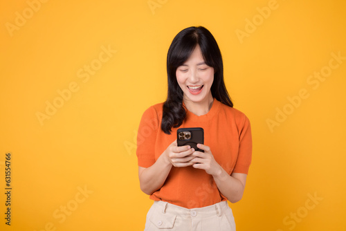 Happy Asian woman in her 30s using smartphone, wearing orange shirt, on vibrant yellow background. Mobile tech concept.