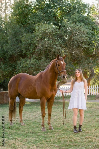 Young girl wearing a white dress and holding a leash of the brown horse next to her