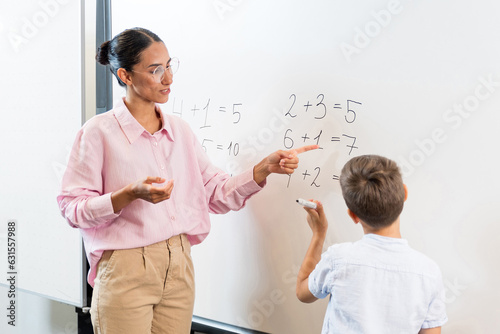 Happy schoolteacher helping a schoolboy doing an addition on a blackboard. Mixed race female teacher in glasses explaining math examples to schoolboy. Communication and discussion about learning