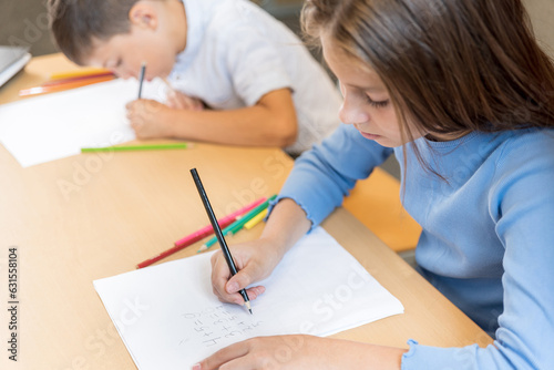 Two primary school pupils at their desks in class, close up. A schoolgirl and a schoolboy leaned over notebooks and write mathematical tasks. The importance of developing correct posture at the desk