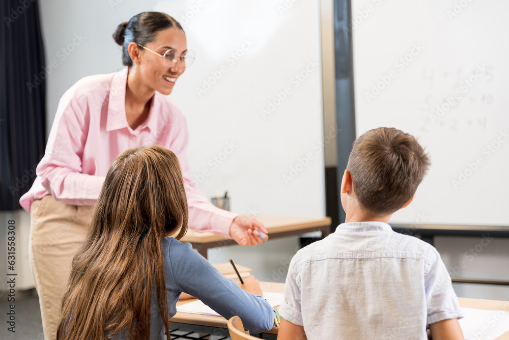 In the foreground, a schoolgirl and a primary school student sit with their backs at their desks. In the background is a mixed race teacher. Young teacher is happy to help children with homework In the foreground, a schoolgirl and a primary school student sit with their backs at their desks. In the background is a mixed race teacher. Young teacher is happy to help children with homework