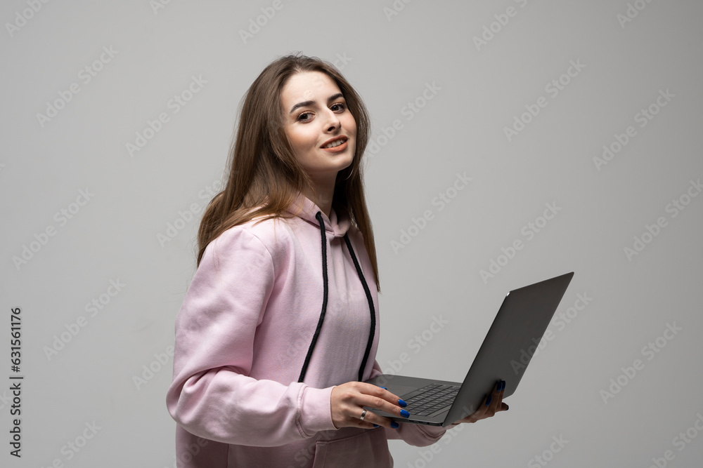 Attractive brunette woman 20s wearing pink hoodie standing working on laptop pc computer isolated on grey wall background studio portrait. Attractive brunette woman 20s wearing pink hoodie standing working on laptop pc computer isolated on grey wall background studio portrait.