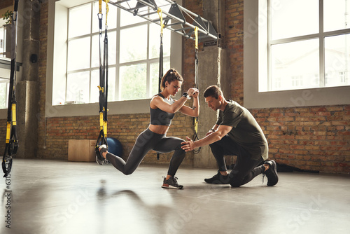 Doing squat exercise. Confident young personal trainer is showing slim athletic woman how to do squats with Trx fitness straps while training at gym.