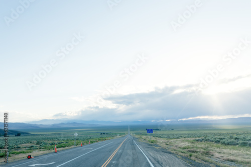 A long asphalted straight road stretches far into the horizon, on which blue mountains can be seen. The rays of the sun break through the large clouds over the road. blue sky before sun © Liudmila