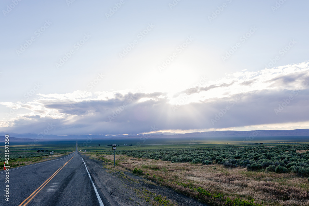 A long asphalted straight road stretches far into the horizon, on which blue mountains can be seen. The rays of the sun break through the large clouds over the road. blue sky before sun