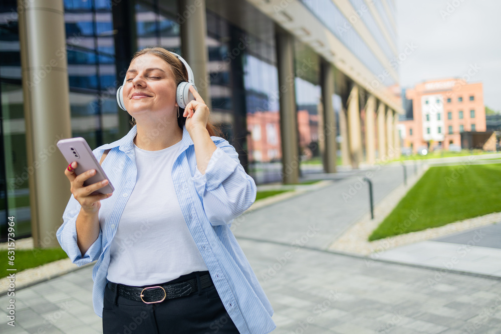 Fototapeta premium Young happy woman wearing suit using wireless earphones holding smartphone listening to music and singing, walking on urban city street. High quality photo