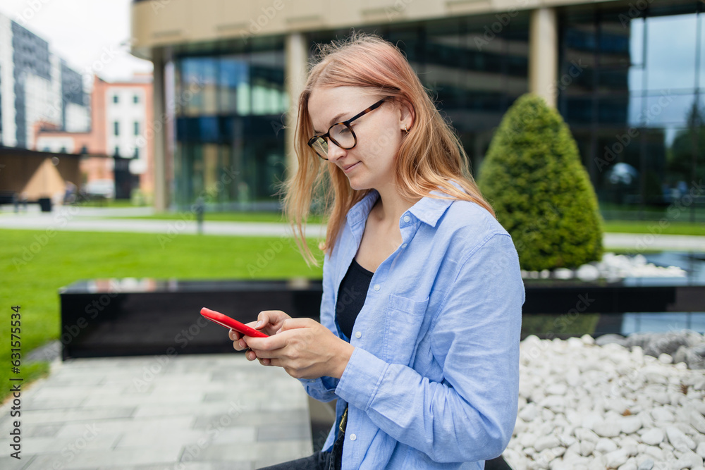 Fototapeta premium Cheerful happy stylish woman using smartphone online app while sitting near the mall. Happy woman in a blue shirt walks down the street looking at her smartphone. An exciting application used for