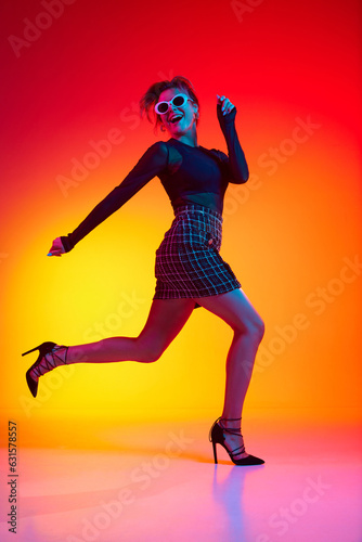 Full lenght photo of running woman wearing fashionable with sunglasses in transparent top and skirt posing over gradient yellow-red neon background.