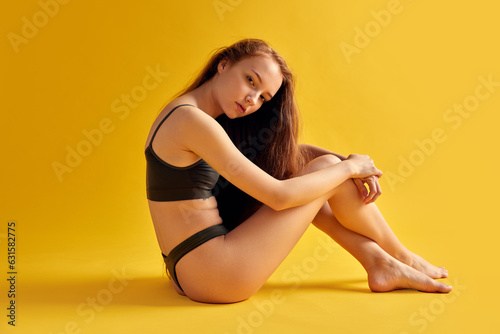 Side view of tender young girl with slim body, sitting and put her head on knees looking at camera in black underwear against bright background.