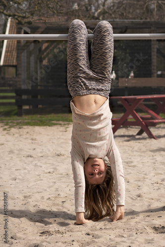 Girl child hanging upside down on a bar in the playground touching the sand with her hands in summer or spring. Outside in the playground. 