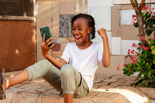 Fotografi A young African girl sitting on the ground looking at her cell phone