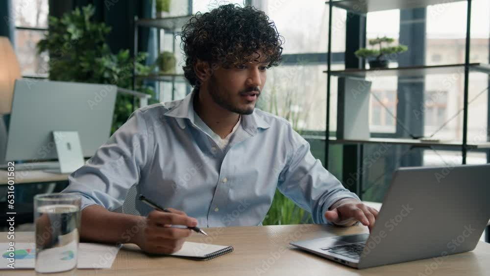 Arabian Indian latin man businessman guy write business notes ideas in notebook managing corporate tasks writing schedule with pen on desk male worker employee working with laptop computer in office