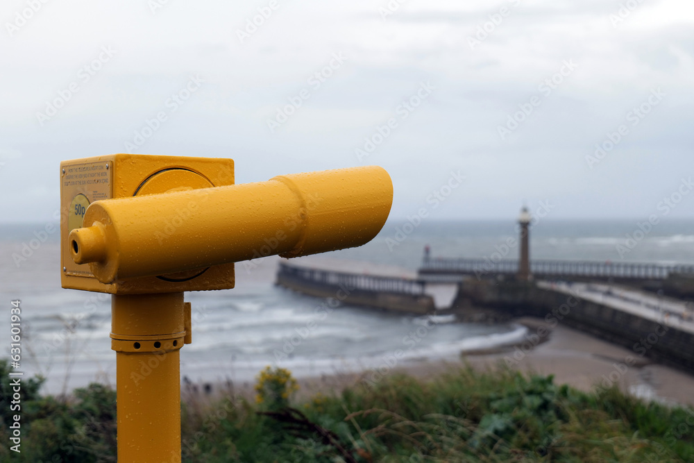 A yellow tourist telescope looks out over the bay of the seaside town of Whitby, UK. Its been raining and drops or water can be seen on it. A yellow tourist telescope looks out over the bay of the seaside town of Whitby, UK. Its been raining and drops or water can be seen on it.