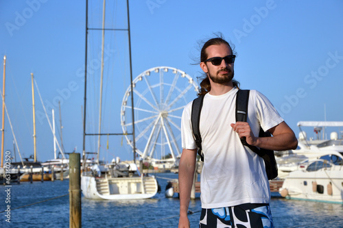 Young caucasian man with long hair standing on a port pier wearing casual beach clothing and a bagpack on a sunny summer day.  photo