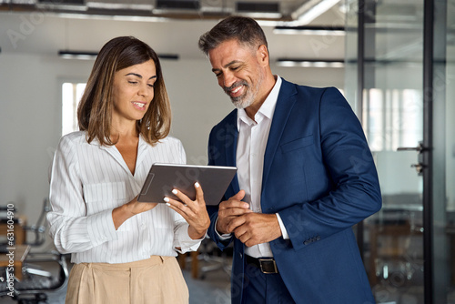 Two diverse partners walking mature Latin business man and European business woman discussing project on tablet in office. Two colleagues of professional business people working together at workspace.