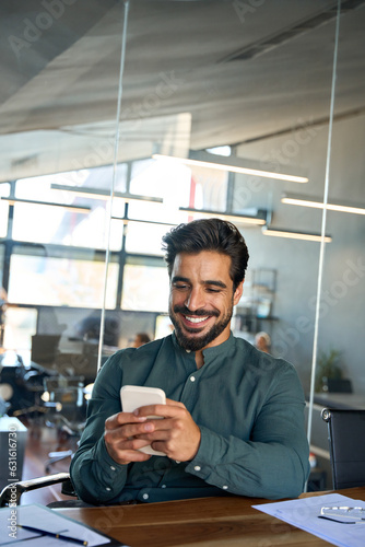 Smiling young Latin business man executive, businessman employee sitting at desk in office holding smartphone using mobile cell phone managing digital apps on cellphone at work. Vertical